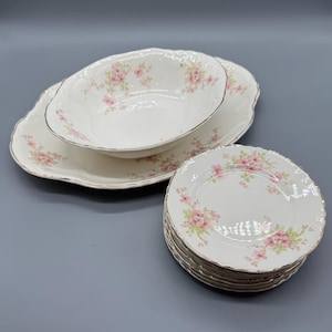 May include: A set of white porcelain tableware featuring a pink floral design and gold edging. The collection includes a serving platter, a bowl, and a stack of plates. The items are displayed on a grey surface.