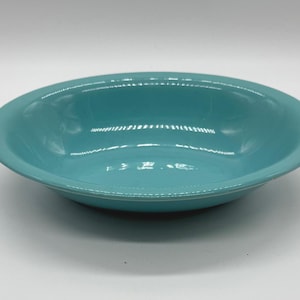 May include: A turquoise ceramic bowl with a glossy finish. The bowl has a wide, shallow design with a slightly raised rim. The interior and exterior are the same color, and the bowl sits on a white surface.