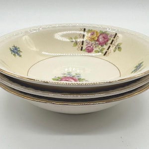 May include: A stack of four cream-colored ceramic bowls with gold rims. Each bowl features a floral design with pink, yellow, and blue flowers. The bowls have a decorative edge.