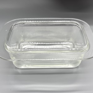 May include: Clear glass rectangular baking dish with a matching lid. The dish has a ribbed texture and rounded corners. The lid fits snugly over the dish, creating a tight seal. The dish is designed for baking and serving.