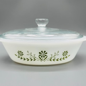 May include: A round white milk glass casserole dish with a clear glass lid featuring a knob handle. The exterior of the dish is decorated with a repeating green floral pattern consisting of stylised daisies and leafy sprigs around the centre.