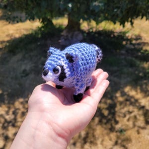 May include: A small, handcrafted crochet raccoon toy made with light blue yarn. It features black yarn accents around the eyes, black ears, and black paws. The raccoon has large, round, dark plastic safety eyes and a small white muzzle area.