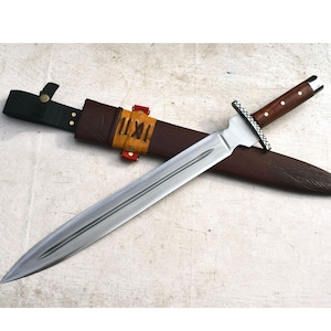May include: A long, silver blade sword with a brown wooden handle and a leather sheath. The handle has a dark finish with a decorative metal guard. The sheath is brown leather with a yellow strap and red accents.