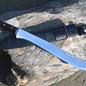 May include: A large, silver-bladed machete with a dark brown wooden handle and a black leather sheath. The blade has several circular cutouts. The machete rests on a weathered wooden surface.