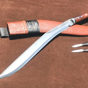 May include: A large, curved blade knife with a wooden handle and two smaller knives, all with silver blades. The knives are displayed with a brown leather sheath and a brown background.
