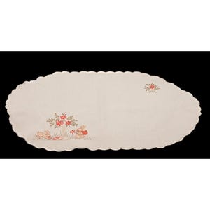 May include: Oval off-white Easter table runner with a scalloped edge. Embroidered with two bunnies, flowers, and eggs in shades of red, orange, and green. The runner is made of a soft, woven fabric.