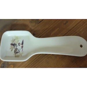 May include: A white ceramic spoon rest with a bird and floral design. The rest has a wide, rectangular area for the spoon and a long handle with a hole for hanging. The bird design features two birds and foliage in shades of gray, brown, and green.