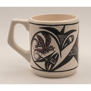 May include: Cream-colored ceramic mug with a square handle. The mug features a black and burgundy bird and floral design on a white background. The mug has a black border around the top and bottom.