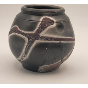 May include: A small, round, dark gray ceramic pot with a textured, cross-shaped design in purple and white. The pot has a dark rim and a small, circular indentation on the side. The design is slightly raised, adding texture to the surface.
