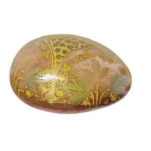 May include: An oval-shaped decorative box with a pinkish-beige base color. The box is adorned with intricate gold and green floral and tree-like patterns. The design is detailed and appears to be hand-painted, adding to its unique charm.