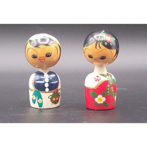 May include: Two vintage wooden dolls with painted faces and traditional clothing. One doll wears a blue vest over a white shirt with a floral design, the other a red and white dress with floral accents. The dolls have black hair and are on a dark background.