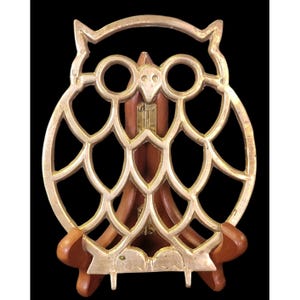 May include: A decorative owl-shaped book stand. The stand features a gold-colored metal owl design with cut-out details, mounted on a wooden base. The owl's eyes and facial features are clearly defined. The wooden base is a warm brown color.