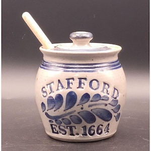 May include: A stoneware jar with a lid and a wooden spoon. The jar is off-white with blue accents and the word "STAFFORD" and "EST. 1664" in blue. The jar has a blue floral design.