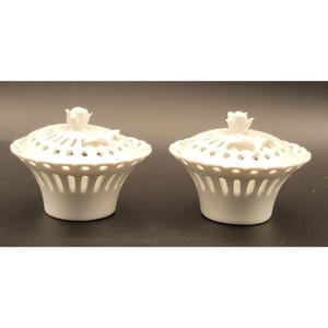 May include: Two white porcelain lidded containers. Each container has a pierced design on the lid and body, with a rose-shaped finial on top. The containers are on a dark surface.