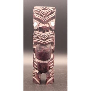 May include: A carved wooden tiki statue with a dark reddish-brown finish. The statue features intricate carvings, including facial features, geometric patterns, and a stylized body. White accents highlight the eyes. The base has an opening.