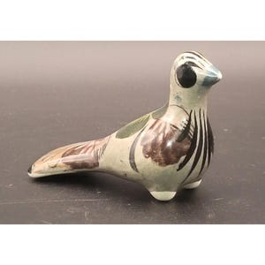 May include: A ceramic bird figurine with a light gray base color, featuring brown, green, and black painted details. The bird has a long tail and a small beak, with a black eye and a blue tip on the beak. The figurine is resting on three small feet.