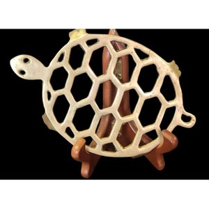 May include: A decorative brass turtle-shaped trivet with a honeycomb design. The trivet is supported by a wooden stand. The turtle has a head, tail, and flippers. The trivet is approximately 8 inches in diameter.
