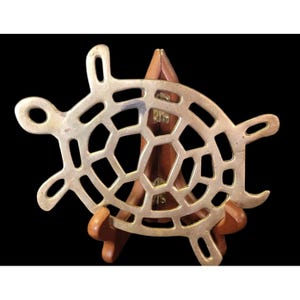 May include: A brass-colored metal trivet in the shape of a turtle. The trivet has a geometric pattern within the turtle's shell and is supported by a wooden stand. The background is black.