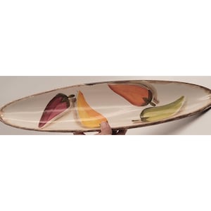 May include: An oval ceramic serving platter with a cream-colored interior and a brown rim. The platter is decorated with hand-painted chili peppers in shades of red, orange, yellow, and green. A hand is holding the platter.