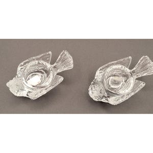 May include: Two clear glass fish-shaped candle holders. Each holder has a round indentation for a candle. The fish have textured details on their fins and bodies. The candle holders are on a gray surface.