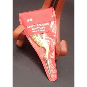 May include: Gold-colored stork scissors in a red plastic sleeve. The scissors are in the shape of a stork with a detailed design. The sleeve has the text "STORK SCISSORS 27-07024 MADE IN JAPAN".
