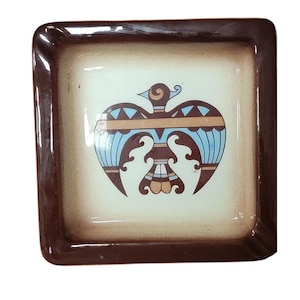 May include: Square ceramic dish with a brown border and a cream-colored center. The center features a stylized bird design in blue, brown, and orange. The dish is likely used for serving or display.