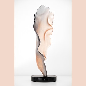 May include: A glass sculpture with a flowing, organic shape. The sculpture is a gradient of colors, from a dark gray at the top to a light peach at the bottom. The sculpture is mounted on a black base.