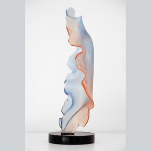 Può includere: Scultura astratta in vetro con una forma organica e fluida. La scultura è realizzata in vetro trasparente con sfumature blu e arancioni. È esposta su una base nera.