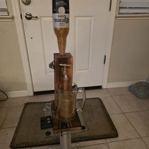 May include: A custom beer tap made from a Corona Extra bottle. The tap is mounted on a wooden stand with a metal base, and a glass mug is positioned below to catch the beer. The tap is open, and beer is flowing into the mug.