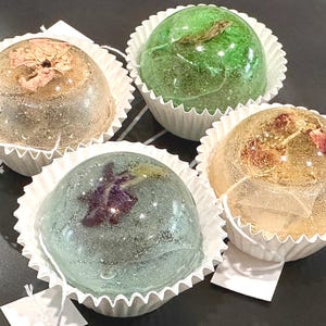 May include: Four clear glass spheres with dried flowers inside, each sphere is in a white paper cupcake liner. The flowers are different colors, including green, brown, and purple.