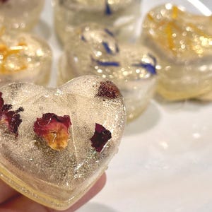 May include: Heart-shaped, clear resin objects with embedded dried flowers. The resin is slightly yellowed, and the flowers are red, orange, and blue. Several of these decorative pieces are visible, some in focus, others blurred.