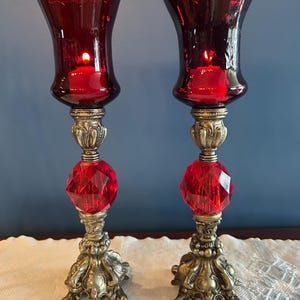以下が含まれることがあります： ルビーレッドのガラスシェードとファセットクリスタルアクセントが付いた、2つの装飾的な燭台。燭台は、複雑なディテールと点灯した赤いろうそくが付いた、銀色の金属ベースを備えています。