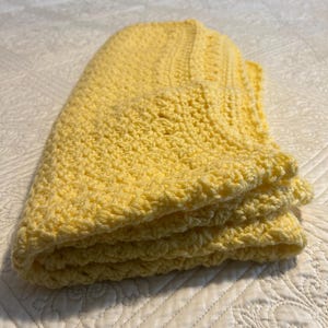May include: A folded, handmade yellow crochet blanket. The blanket is made of soft yarn and has a textured surface. The blanket is folded neatly, showing the layers of the crochet work. The colour is a bright, sunny yellow.