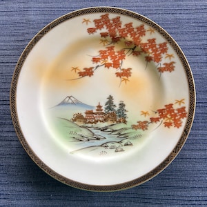 Puede incluir: Un plato de cerámica blanca con un borde dorado y un diseño de inspiración japonesa. El plato presenta una montaña, un río, una pagoda y un árbol con hojas rojas.