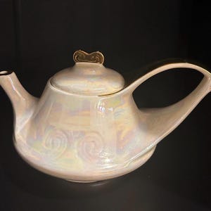 May include: Iridescent white ceramic teapot with a gold handle and accents. The teapot has a curved spout and a lid with a decorative gold finial. The body of the teapot has a pearlescent finish, reflecting a range of colours.