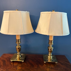 Puede incluir: Un par de lámparas de mesa con bases de latón pulido, plataformas cuadradas y columnas escalonadas. Cada lámpara tiene una pantalla de tela color crema con un diseño estructurado de paneles y un pequeño remate de latón en la parte superior.