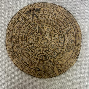 May include: A circular, tan-colored artifact with intricate carvings. The design features concentric circles filled with symbols and glyphs, possibly representing a calendar or astronomical chart. The artifact has a small hole at the top.