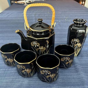 May include: A black ceramic tea set with gold trim and floral designs. The set includes a teapot with a woven handle, a vase, and four cups. The tea set is displayed on a blue tablecloth.