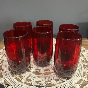 May include: Six red-tinted drinking glasses are arranged on a white lace doily. The glasses are cylindrical with a slightly tapered shape. The red color is consistent throughout the glasses, and they appear to be made of glass.