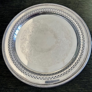 Op de afbeelding: Een ronde, zilverkleurige serveerschaal met een sierlijk, reliëf bloemmotief in het midden. De schaal heeft een decoratieve opengewerkte rand en een touwachtige rand. De schaal is van metaal en reflecteert licht.
