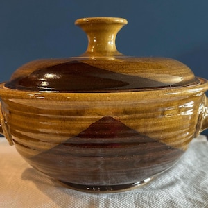 Puede incluir: Una fuente de cerámica cubierta con tapa y dos asas. La fuente está esmaltada en tonos marrones y beige, con un patrón geométrico en la parte delantera. La tapa tiene un pequeño pomo en la parte superior.