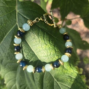 May include: A beaded bracelet featuring light blue and navy blue beads, interspersed with gold-colored spacers. The bracelet has a gold-colored toggle clasp. The jewelry is displayed on a large green leaf.