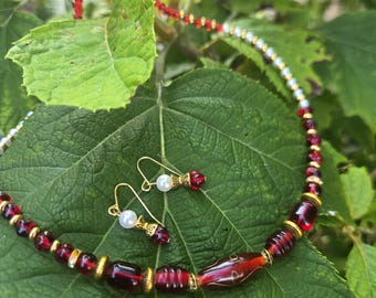 May include: A handmade jewelry set featuring a necklace and matching earrings. The necklace and earrings have red and gold beads, with a pearl accent on the earrings. The jewelry is displayed on a large green leaf.