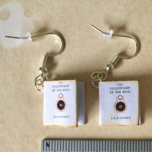 Può includere: Orecchini e un piccolo libro, entrambi con la copertina di "The Fellowship of the Ring" di J.R.R. Tolkien. Gli orecchini hanno ganci argentati. L'insieme ha un aspetto vintage.