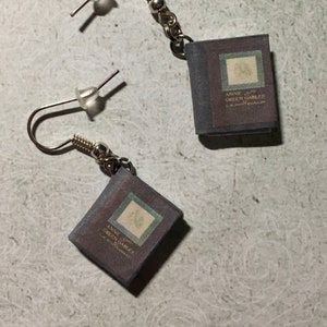 Può includere: Orecchini pendenti con ciondoli a forma di libro in miniatura. I ciondoli a forma di libro sono viola scuro con un design verde e bianco e il testo "Anne of Green Gables". Gli orecchini hanno ganci color argento e piccole perline.