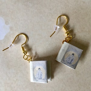 Può includere: Un paio di orecchini fatti a mano con ciondoli a forma di libro in miniatura. Ogni ciondolo a forma di libro mostra una figura seduta con un'aureola dorata. Gli orecchini hanno ganci per le orecchie color oro e piccoli anelli color oro.