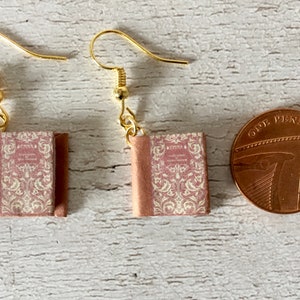 Può includere: Orecchini dorati a forma di libro con copertina a fantasia rosa e bianca. Gli orecchini sono a forma di libri in miniatura, con un gancio dorato per orecchie forate. Una moneta è accanto.