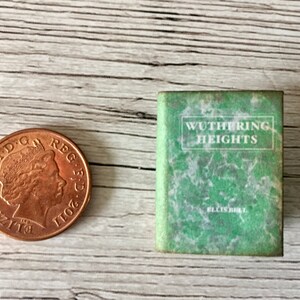 May include: A miniature book titled "Wuthering Heights" with a green cover and white text, next to a copper penny. The book's cover has a marbled effect. The penny is a United Kingdom one penny coin.