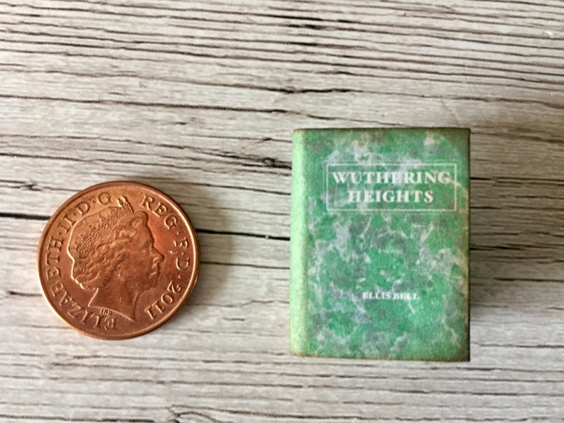 May include: A miniature book titled "Wuthering Heights" with a green cover and white text, next to a copper penny. The book's cover has a marbled effect. The penny is a United Kingdom one penny coin.