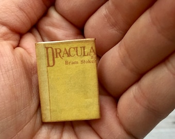 Pode incluir: Um livro em miniatura com capa amarela e o título "DRACULA" de Bram Stoker em letras vermelhas. O pequeno livro é segurado em uma mão, mostrando seu tamanho e detalhes.